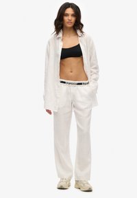Witte linnen overhemd met knopen, zwarte sport-bh, witte broek met merk tailleband en beige sneakers. Casual en loose-fit ontwerp.