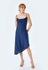 Abito blu in satin con scollo a cowl, spalline sottili e orlo asimmetrico. Il tessuto ha una consistenza liscia che esalta la caduta del capo.
