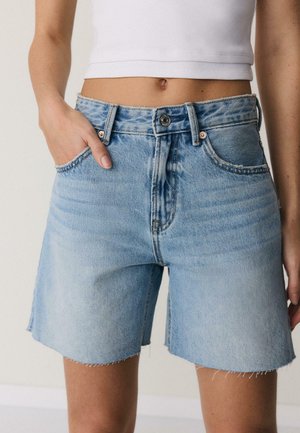 Frau trägt hellblaue Jeansshorts und ein weißes, bauchfreies Tanktop, mit linker Hand in der Tasche vor einfarbigem Hintergrund.