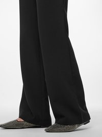 Pantalons larges noirs associés à des chaussures à bout pointu ornées de clous en argent, présentant une texture élégante et un design à talon plat.