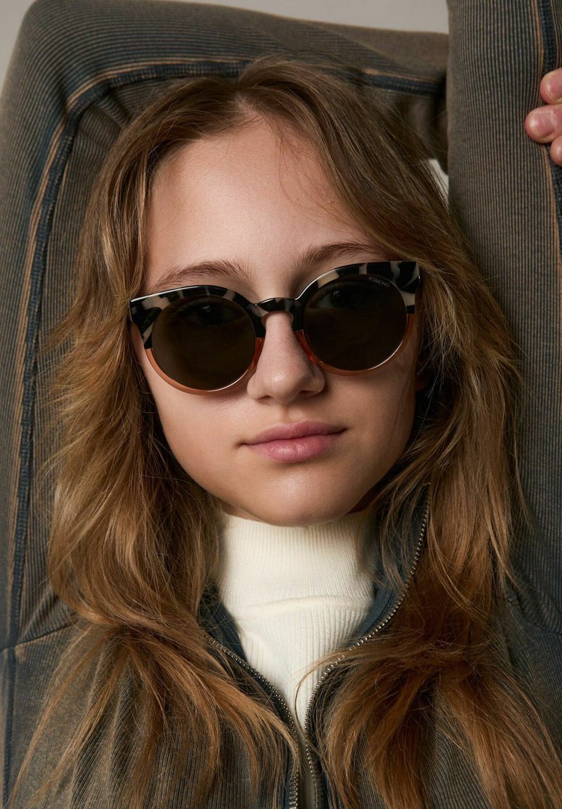 Gafas de sol con un audaz patrón geométrico en blanco y negro, lentes oscuros redondos y una línea de cejas acentuada, llevadas con un cuello de tortuga ligero.