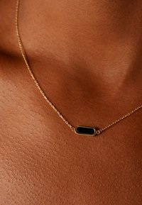 Gouden ketting met een ovaal zwarte hanger. Gladde textuur en een fijn ontwerp, rustend op een lichtbruine huidtint.
