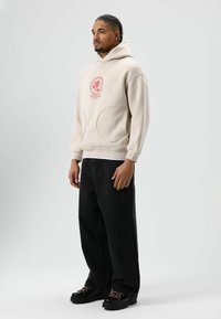 Beige hoodie met rode grafische print, grote voorzakken, losvallende pasvorm. Gecombineerd met zwarte wijde broekspijpen en zwarte chunky schoenen.