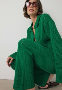 Zaļš adīts kardigāns un atbilstošas bikses ar brīvu piegriezumu. Iespraugenās pogas un vaļējo austs tekstūra. Modelis valkā tumšas saulesbrilles.