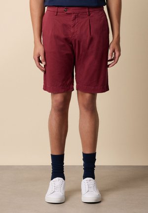 Uomo con pantaloncini rossi al ginocchio, calzini blu navy e scarpe da ginnastica bianche in piedi su uno sfondo neutro.