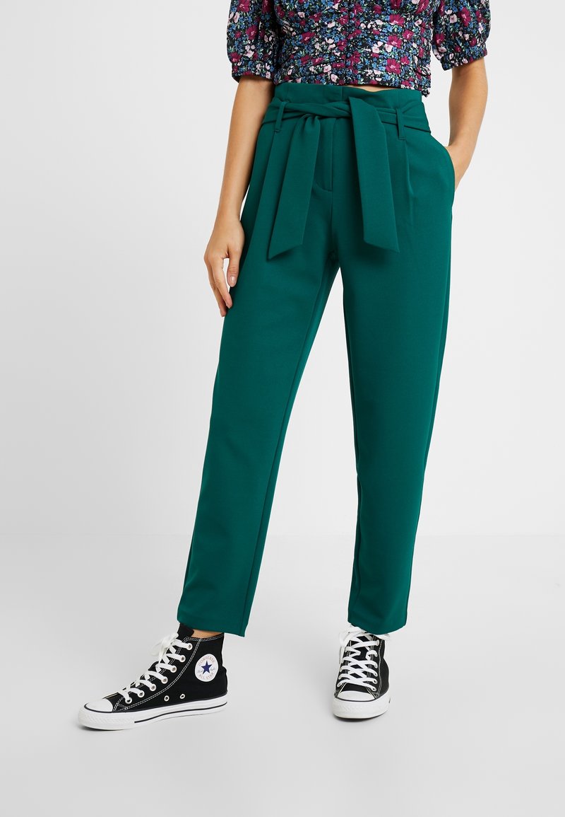 Femme portant un pantalon taille haute couleur sarcelle avec une large ceinture nouée et des baskets Converse noires montantes, debout avec une main dans la poche.