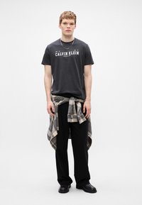 Mustvalge puuvillane T-särk valge "CALVIN KLEIN" logoga, koos lõdva mustade pükstega ja plüüšist kattega särgiga, mis on seotud talje ümber. Mustad kingad.