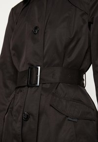 Ciemnobrązowy płaszcz trenczowy z paskiem, podwójnym zapięciem, dużymi guzikami oraz bocznymi kieszeniami, wykonany z gładkiego, lekkiego materiału.