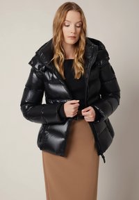 Czarna puchowa kurtka o błyszczącej, pikowanej powierzchni, z kapturem i zapięciem na zamek. Stylizowana z czarną bluzką i brązową spódnicą. Prosty, elegancki design.