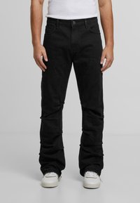 Muž oblečený v čiernych cargo nohaviciach a bielych teniskách stojí na jednoduchom pozadí, ruky uvoľnene pri bokoch.