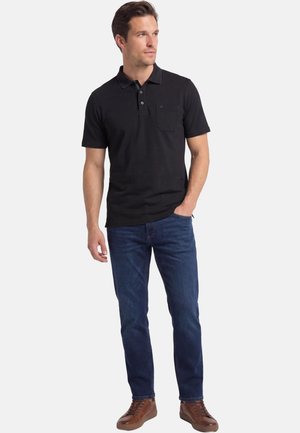 Mann trägt schwarzes kurzärmeliges Poloshirt, blaue Jeans und braune Sneaker, steht mit einer Hand in der Tasche und schaut zur Seite.