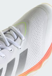 Chaussure de sport blanche avec dessus en mesh, lacets blancs, rayures grises, accent vert néon et bord de semelle orange sur fond clair.