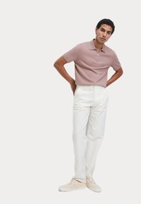 Kortærmet polo med lynlås i lyserød, parret med hvide bukser; skoene er beige. Modellen står med hænderne på hofterne.