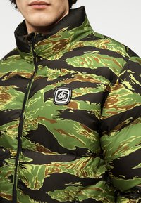 Φουσκωτό μπουφάν σε καμουφλάζ σχέδιο με πράσινα, καφέ και μαύρα χρώματα. Διαθέτει ψηλό γιακά και λογότυπο στο στήθος.