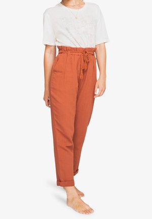 Vrouw die roestoranje, hooggetailleerde broek draagt met een gestrikt riempje en opgerolde boorden, gecombineerd met een los, wit T-shirt dat in de broek is gestopt.