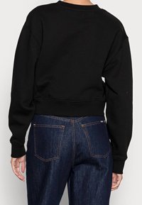 Persona vistiendo una sudadera negra de manga larga corta y jeans de mezclilla azul oscuro de cintura alta, vista desde atrás.