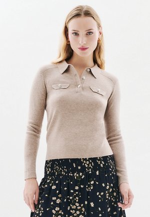 Vrouw met lang blond haar draagt een beige polo trui met lange mouwen en knoopzakken op de borst, gecombineerd met een zwarte bloemetjesrok, staande tegen een witte achtergrond.