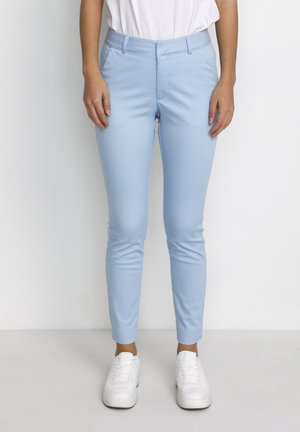 Osoba ubrana w jasnoniebieskie dopasowane spodnie, białe trampki i białą włożoną do środka koszulę, stojąca na tle jednolitego białego tła.