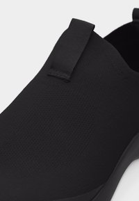 Κοντινό πλάνο μαύρου παπουτσιού χωρίς κορδόνια με υφασμάτινο επάνω μέρος υφής και θηλιά στο πίσω μέρος, πάνω σε λευκό φόντο.