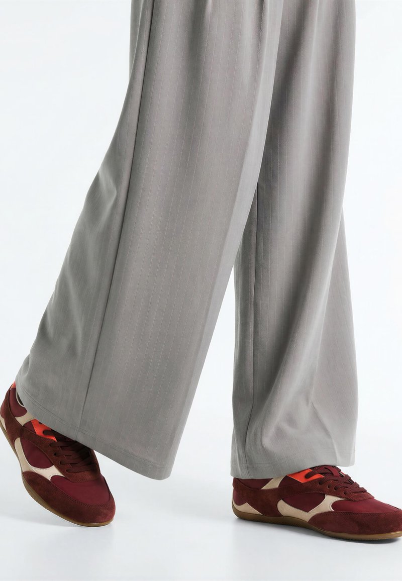 Pantalon large gris clair couvrant les chevilles, associé à des baskets rouges, beiges et orange sur fond blanc.