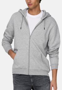 Grijze zip-up hoodie met voorzakken, trekkoordcapuchon en geribbelde manchetten, gedragen over een wit T-shirt, gecombineerd met donkere spijkerbroek.