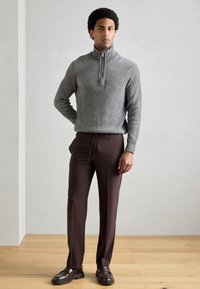 Camisola cinzenta canelada com gola de fecho éclair, combinada com calças castanhas feitas à medida e mocassins escuros, tudo colocado contra um fundo neutro.