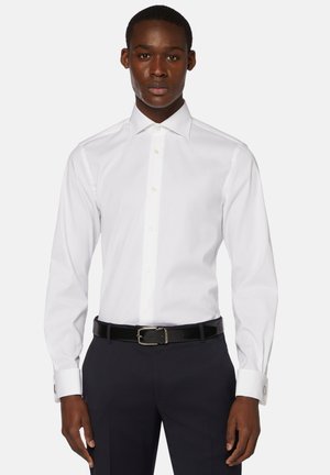 Chemise blanche à manches longues avec col évasé, devant boutonné et détails de poignets doubles, associée à une ceinture noire et un pantalon sombre.