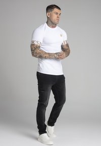 White short-sleeve t-shirt with golden logo, paired with black distressed jeans and white sneakers, showcasing a fitted design.