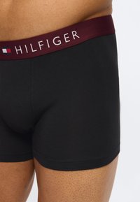 Κοντινό πλάνο ενός ατόμου που φοράει μαύρα μποξέρ με μπορντό ελαστική μέση, με την επωνυμία "Hilfiger" και ένα μικρό λογότυπο με σημαία στην αριστερή πλευρά.