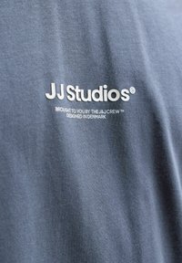 Μπλε ύφασμα με λευκό κείμενο που λέει JJ Studios, προσφερόμενο από The J&J Crew, σχεδιασμένο στη Δανία πάνω σε ένδυμα.