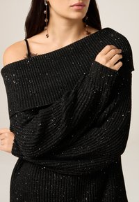 Černý pletený svetr s odhalenými rameny, žebrovanou strukturou a třpytivými detaily. Široké rukávy a přiléhavé manžety. Viditelný průhledný krajkový detail.