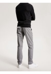 Person som står vänd bort, iklädd en svart långärmad tröja, ljusgrå byxor och vita sneakers mot en enfärgad ljus bakgrund.