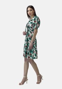 Blumenkleid mit grünen, roten und weißen Mustern, kurzen Puffärmeln, elastischem Bund und knielangem Saum, kombiniert mit weißen Riemchensandalen.