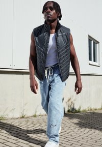 Black quilted sleeveless vest with high collar, white tank top, light blue baggy jeans featuring a Nike logo, and white sneakers.