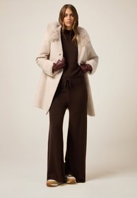 Cappotto beige con un colletto soffice, indossato sopra un abito marrone scuro. Guanti marroni e sneakers con accenti bianchi e beige completano il look.