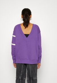 Lila sweatshirt med djup V-rygg, långa ärmar och vita ränder på armarna. Tillverkad av ett mjukt material med en avslappnad passform.