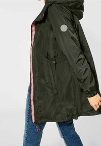 Donkergroene waterdichte jas met capuchon, ritssluiting aan de voorkant, roze accenten en elastische manchetten. Voorzien van een logopatch op de mouw.