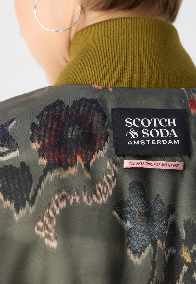 Veste vert olive avec motif floral, col côtelé, et étiquette de marque "Scotch & Soda" à l'arrière. Tissu doux et texturé.