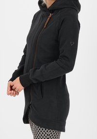 Sudadera negra con capucha, cremallera frontal, mangas largas, bolsillos laterales y un pequeño detalle de corazón en la manga. Tela suave y texturizada.