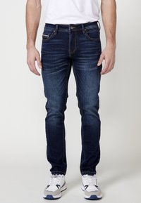 Jeans de mezclilla azul oscuro con un ajuste estrecho, con desvanecimientos sutiles y costuras en contraste. Combinados con zapatillas blancas.