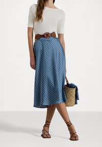 Blå prickig kjol med ett flödande mönster, midikalvlängd, kombinerad med en vit åtsittande topp och ett brunt bälte; accessoriserad med en vävd väska.