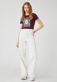 Bordeaux cropped t-shirt med hvid grafik; sat sammen med højtaljede hvide denim flared bukser; modellen står med den ene hånd på hoften i en uformel setting.