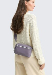 Borsa a tracolla viola in pelle con zip in argento e dettagli a frange, caratterizzata da una forma rettangolare e una superficie texturizzata. Indossata sopra un maglione color crema.