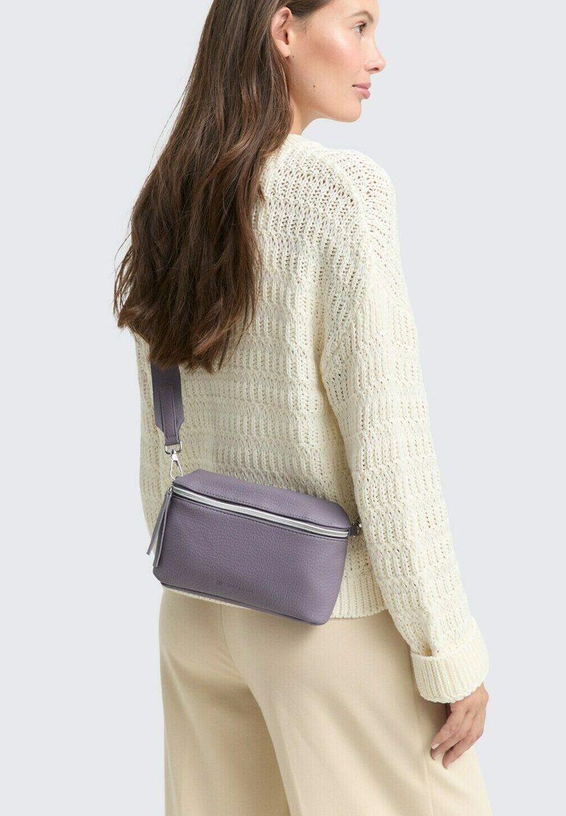 Borsa a tracolla viola in pelle con zip in argento e dettagli a frange, caratterizzata da una forma rettangolare e una superficie texturizzata. Indossata sopra un maglione color crema.