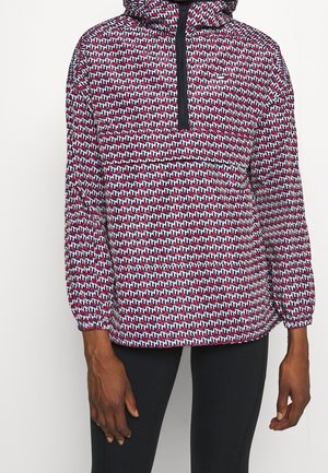 Personne portant un pull à capuche avec un motif géométrique "TH" noir, blanc et rouge et un pantalon noir sur fond uni.