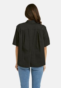 Black short-sleeve shirt with a back pleat design, collar, and a smooth texture. Paired with fitted light blue jeans.