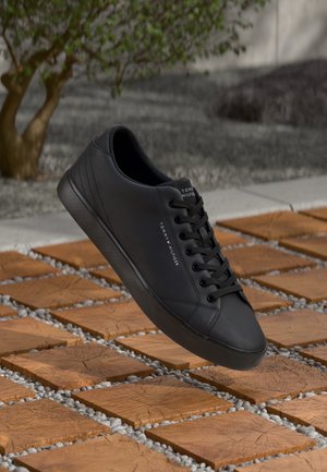 Schwarzer Tommy Hilfiger Sneaker mit Schnürsenkeln, schwebend über quadratischen Holzkacheln und weißen Kieselsteinen, im Freien mit einem Baum im Hintergrund.