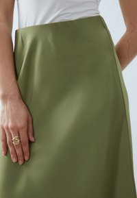 Jupe en tissu lisse vert olive avec une taille haute. Une main repose sur le tissu, portant une bague en or en spirale. Design simple et moderne.
