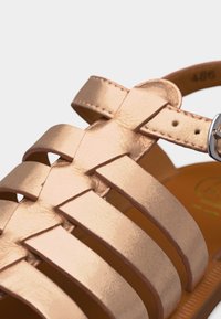 Εγγύς πλάνο ενός δερμάτινου σανδαλιού σε χρώμα ροζ χρυσού με πολλαπλούς ιμάντες και ασημένια αγκράφα σε ταμπά πάτο.