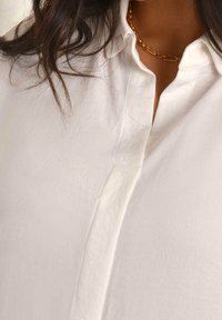 White button-up shirt featuring a collared neckline, soft fabric texture, and a subtle placket detail at the front. Gold chain necklace accent.